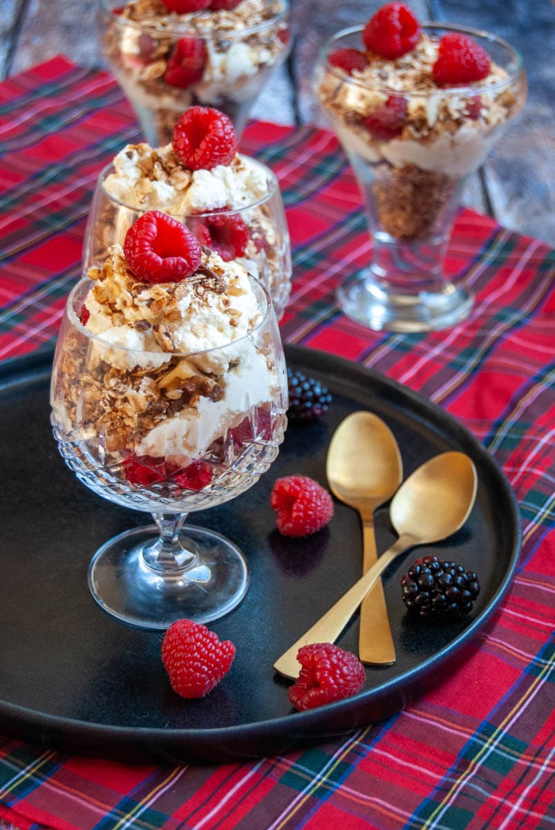 two dessert glasses filled with whipped cream,fresh raspberries and toasted oats on a black plate with two gold spoons and fresh raspberries and a tartan tablecloth.