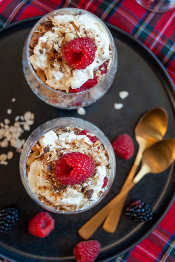 an overhead photograph of two glass dessert dishes filled with Scottish Cranachan - whipped cream, fresh raspberries and toasted oats on a black plate with two gold spoons and a tartan tablecloth. 