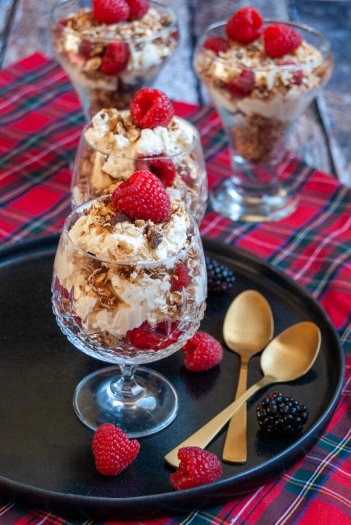 Four dessert glasses filled with whipped double cream, raspberries and toasted oats and nuts on a black plate with two gold serving spoons. 