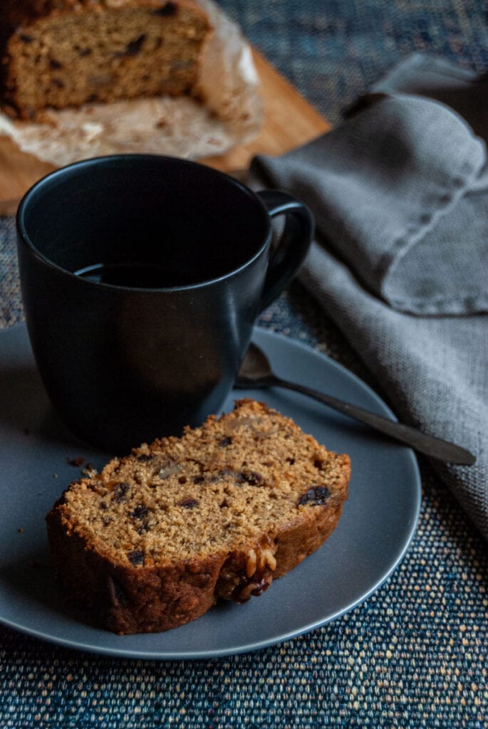 a sliced of date and walnut cake on a grey plate with a black coffee mug and a silver spoon. A grey napkin is sitting beside the plate.