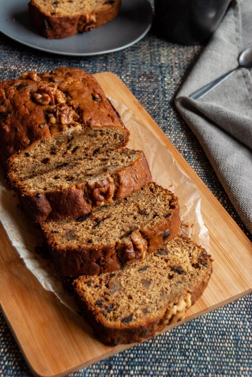 a partially sliced date and walnut cake on a wooden chopping board with a grey napkin, A black coffee mug and a grey plate with a slice of cake can be partially seen in the background.