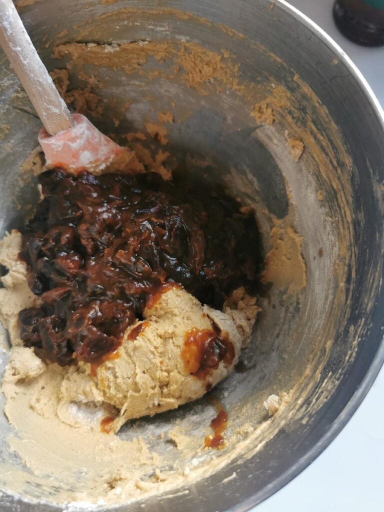 pureed dates being stirred into a silver bowl of cake batter using a wooden handled spatula.