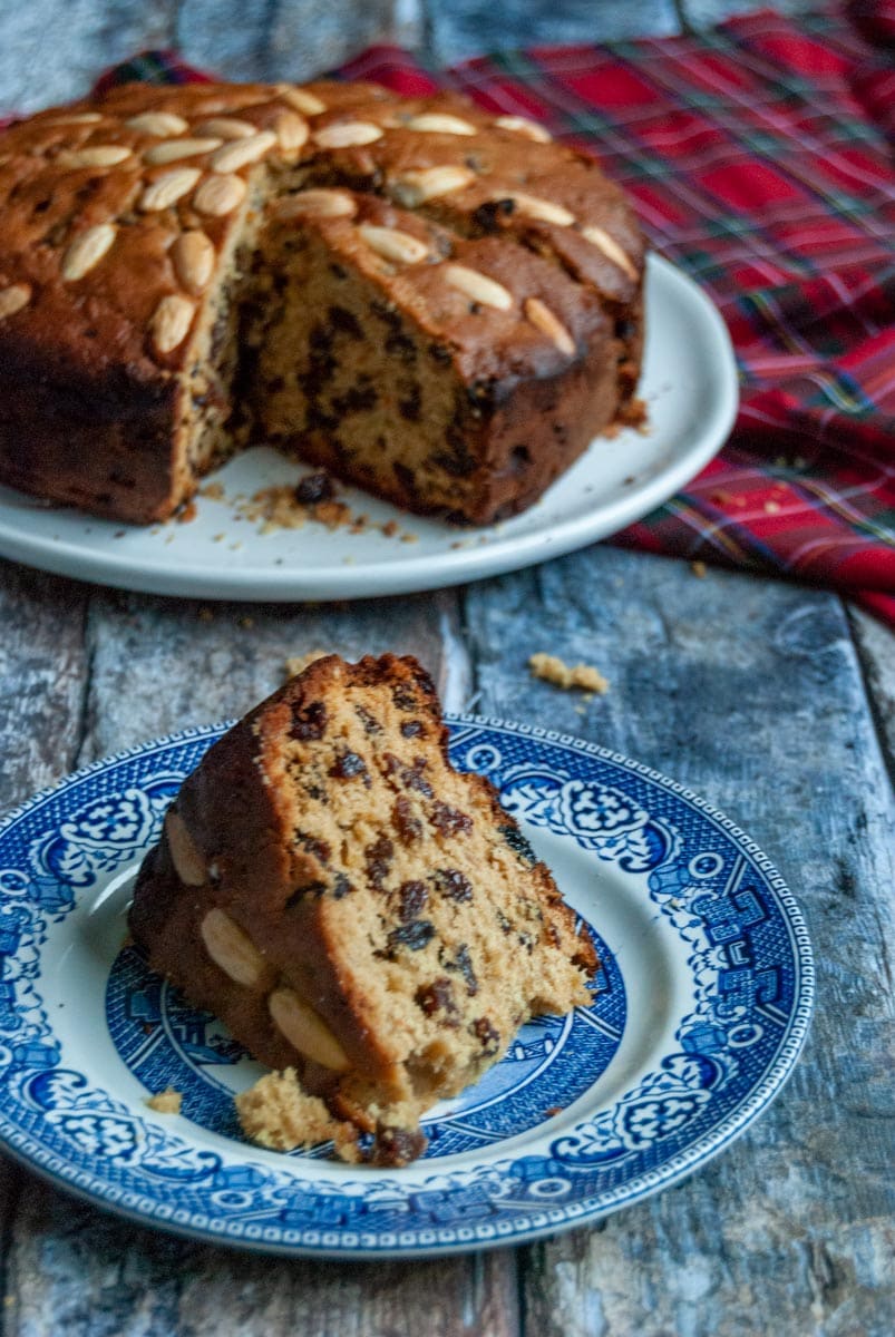 a slice of Dundee fruit cake on a blue and white plate and a large fruit cake sitting on a white plate with a tartan tablecloth.