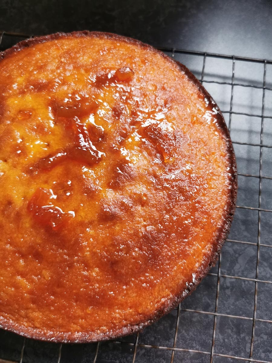 a freshly baked orange cake brushed with marmalade on a black wire cooling rack.