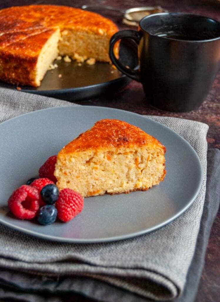 a slice of orange and almond cake with fresh raspberries and blueberries on a grey plate sitting on a grey napkin, a black coffee mug and a large orange cake with a slice removed in the background.