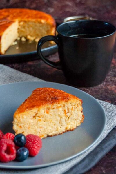 a slice of orange cake with fresh raspberries and blueberries on a blue plate resting on a grey napkin, a black mug of coffee and a large orange cake on a black plate.