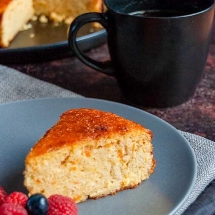 a slice of orange cake with fresh raspberries and blueberries on a blue plate resting on a grey napkin, a black mug of coffee and a large orange cake on a black plate.