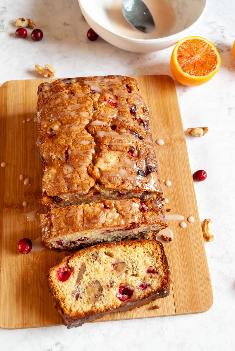 a sliced orange and cranberry loaf cake on a wooden chopping board. A white bowl of orange icing with a silver spoon, fresh cranberries, walnuts and an orange are sitting above the board.