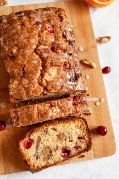 an orange and cranberry loaf cake drizzled with an orange icing glace on a wooden serving board.