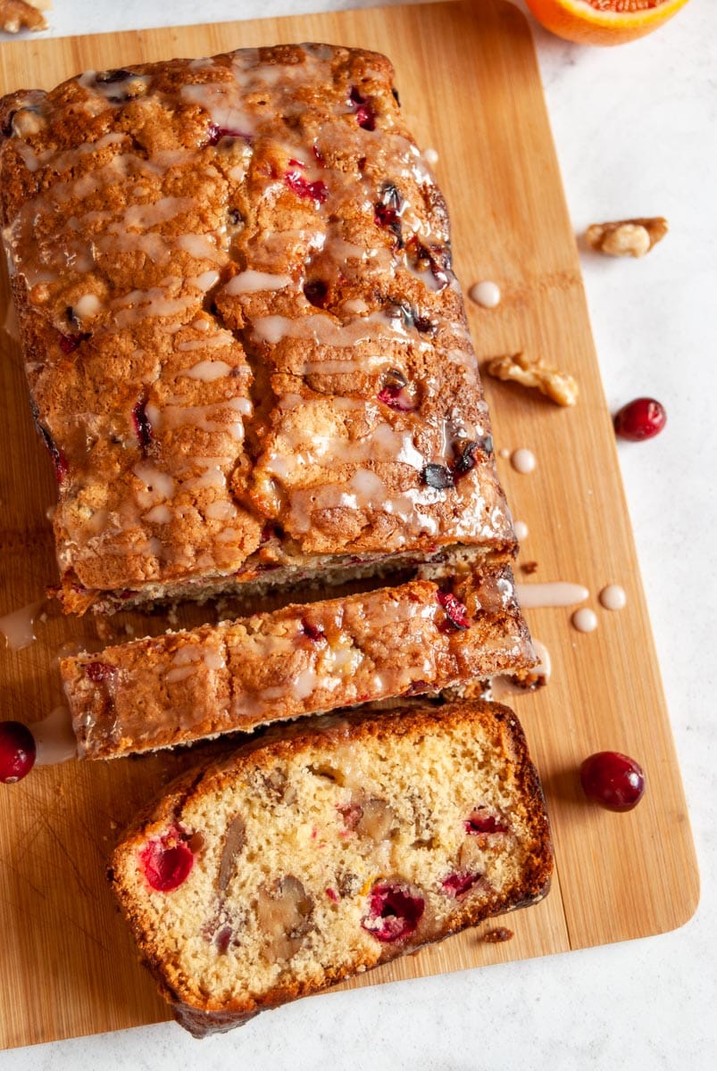 an orange and cranberry loaf cake drizzled with an orange icing glace on a wooden serving board.