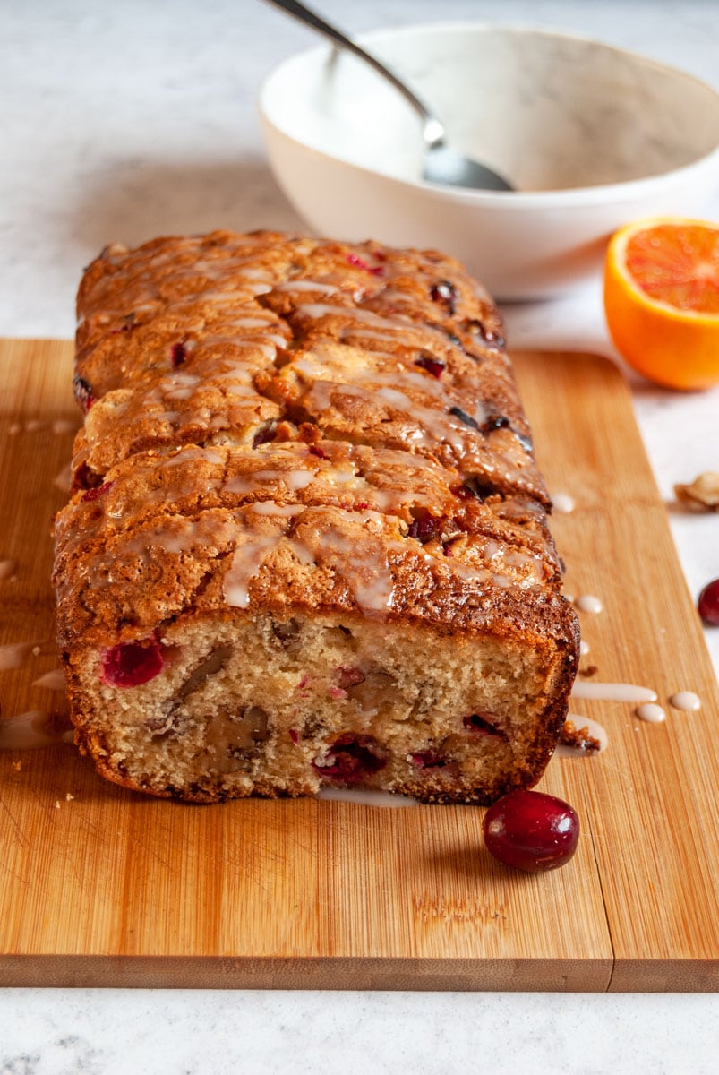 a partially sliced orange and cranberry cake on a wooden chopping board, a white bowl of orange icing with a silver spoon and a sliced orange.