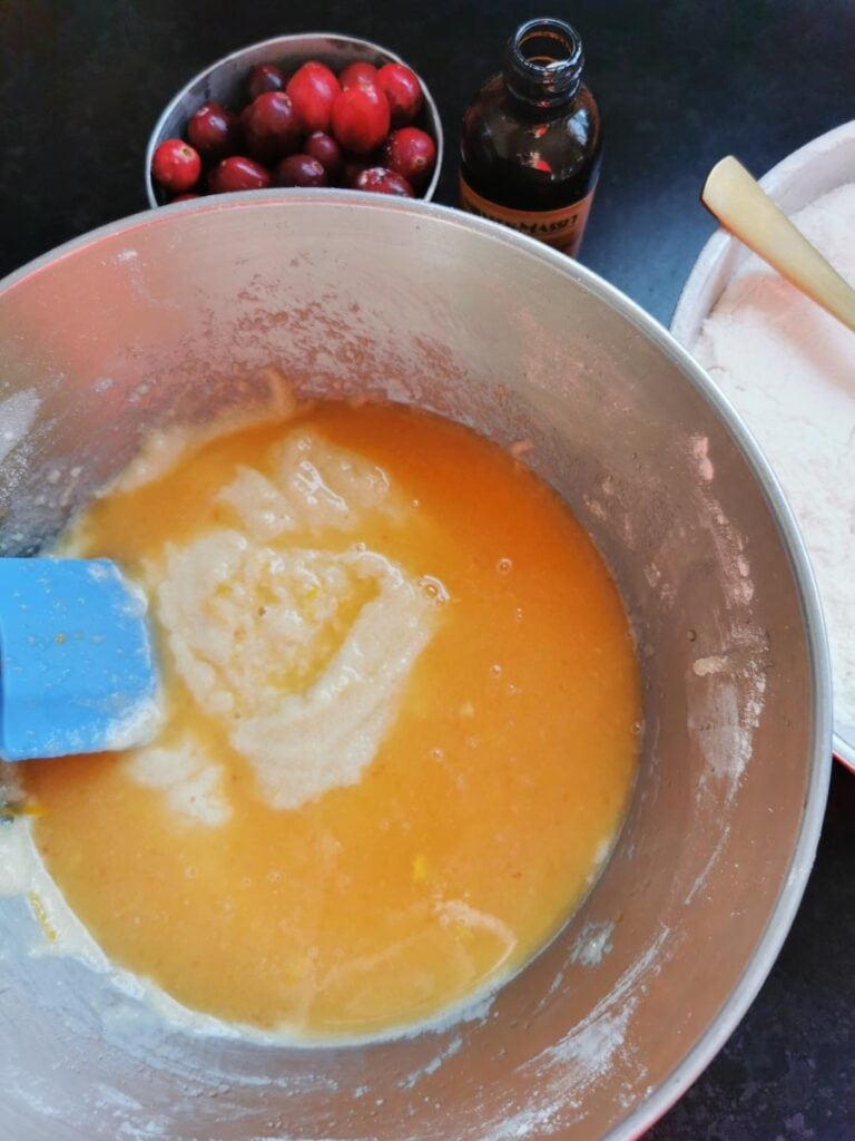 Orange juice being folded into a silver bowl of cake batter with a blue handled spatula.