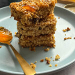 three orange oat bars stacked on top of each other with sultanas and orange marmalade on a blue plate with a gold spoonful of orange marmalade.
