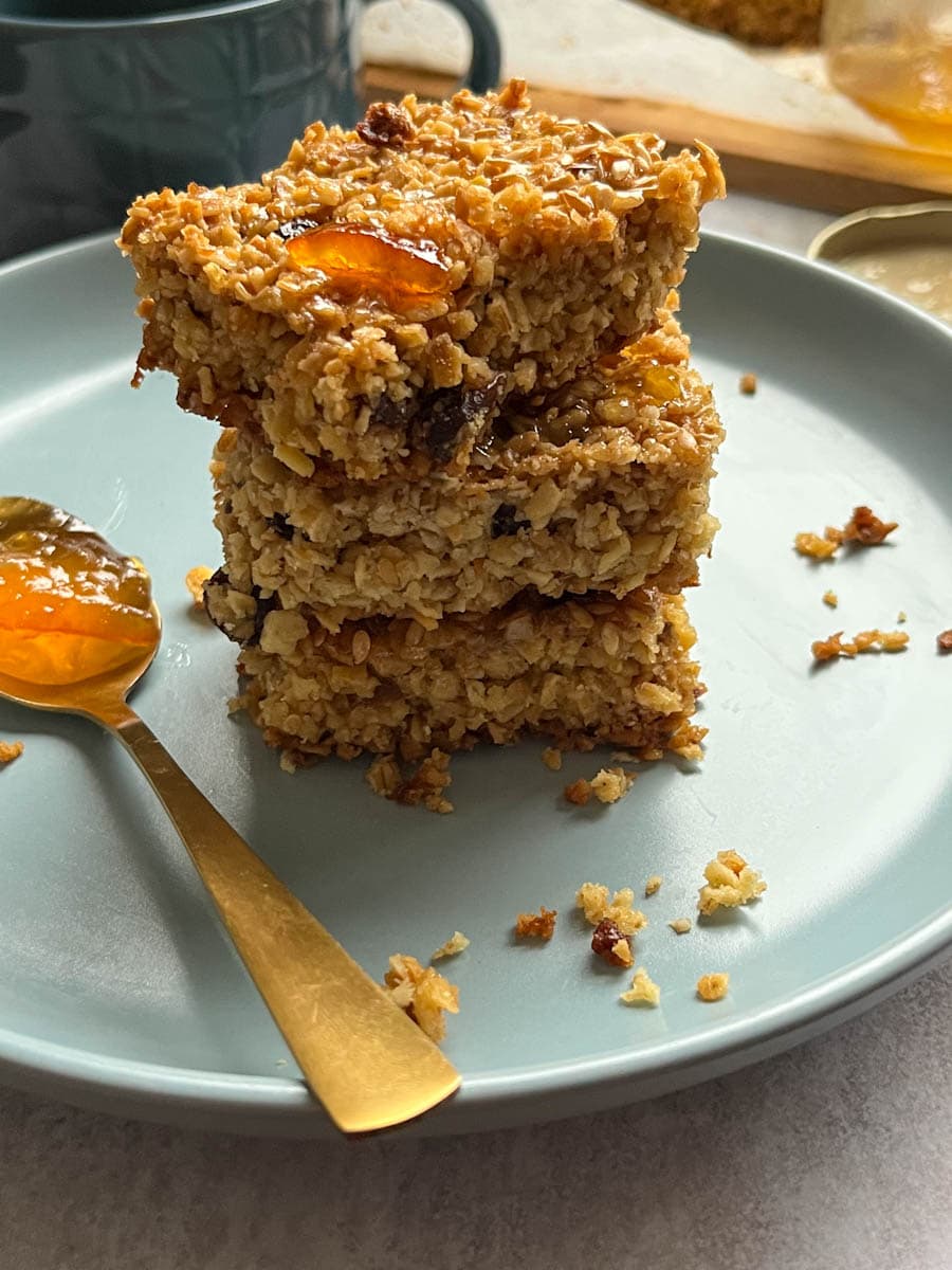 three orange oat bars stacked on top of each other with sultanas and orange marmalade on a blue plate with a gold spoonful of orange marmalade.