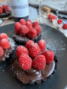 three mini chocolate tarts topped with fresh raspberries on a black plate.