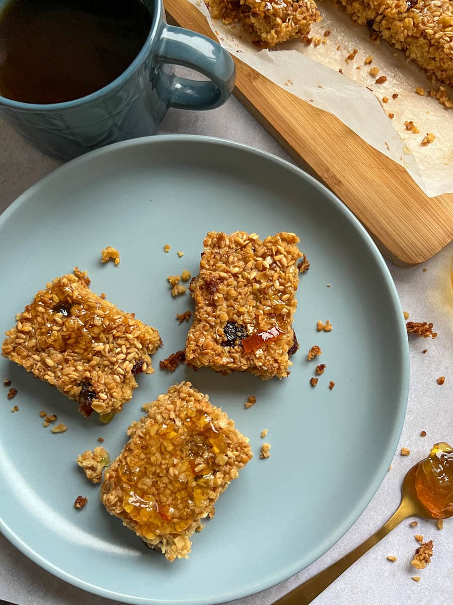 three flapjack oat bars with sultanas and glazed with orange marmalade on a blue plate, a gold spoon with marmalade and a dark blue coffee cup.