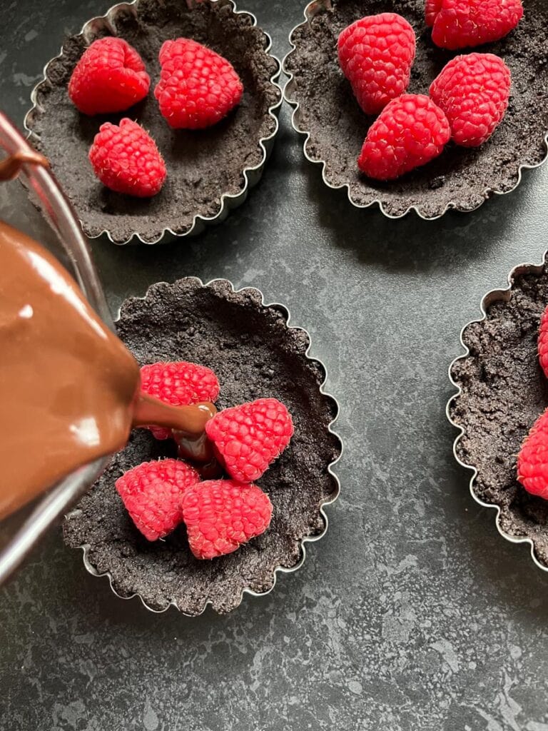 chocolate ganache being poured into four mini tart pans with a chocolate cookie base and filled with fresh raspberries.