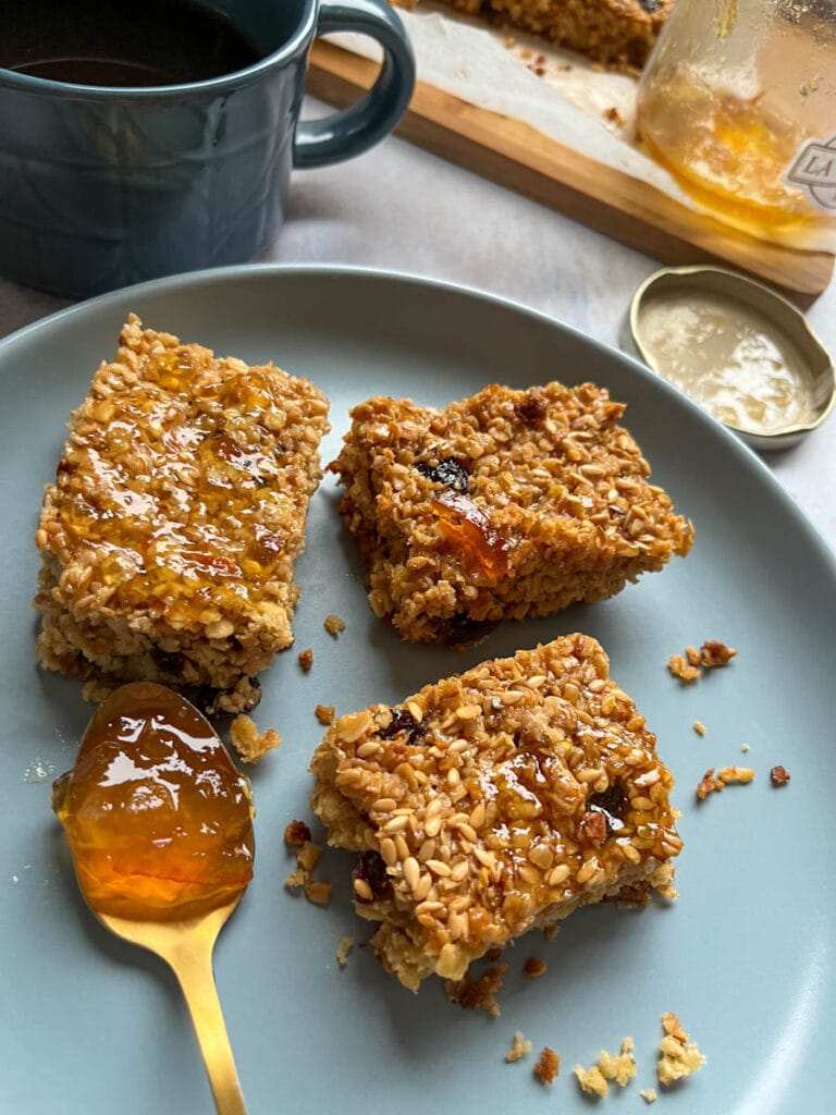 three orange marmalade flapjacks on a blue plate and a gold spoon of marmalade. A dark blue coffee mug and a jar of marmalade can be partially seen in the background.
