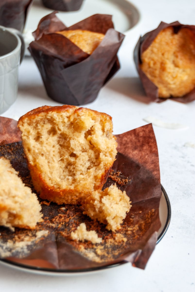 a banana muffin sitting on a brown paper muffin liner. More muffins and a cup of coffee can be partially seen in the background.