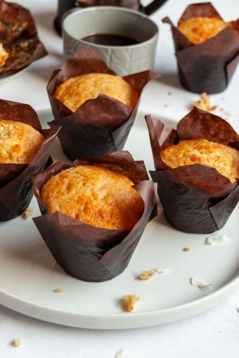 a white plate of banana muffins in brown paper liners with a grey cup of coffee.