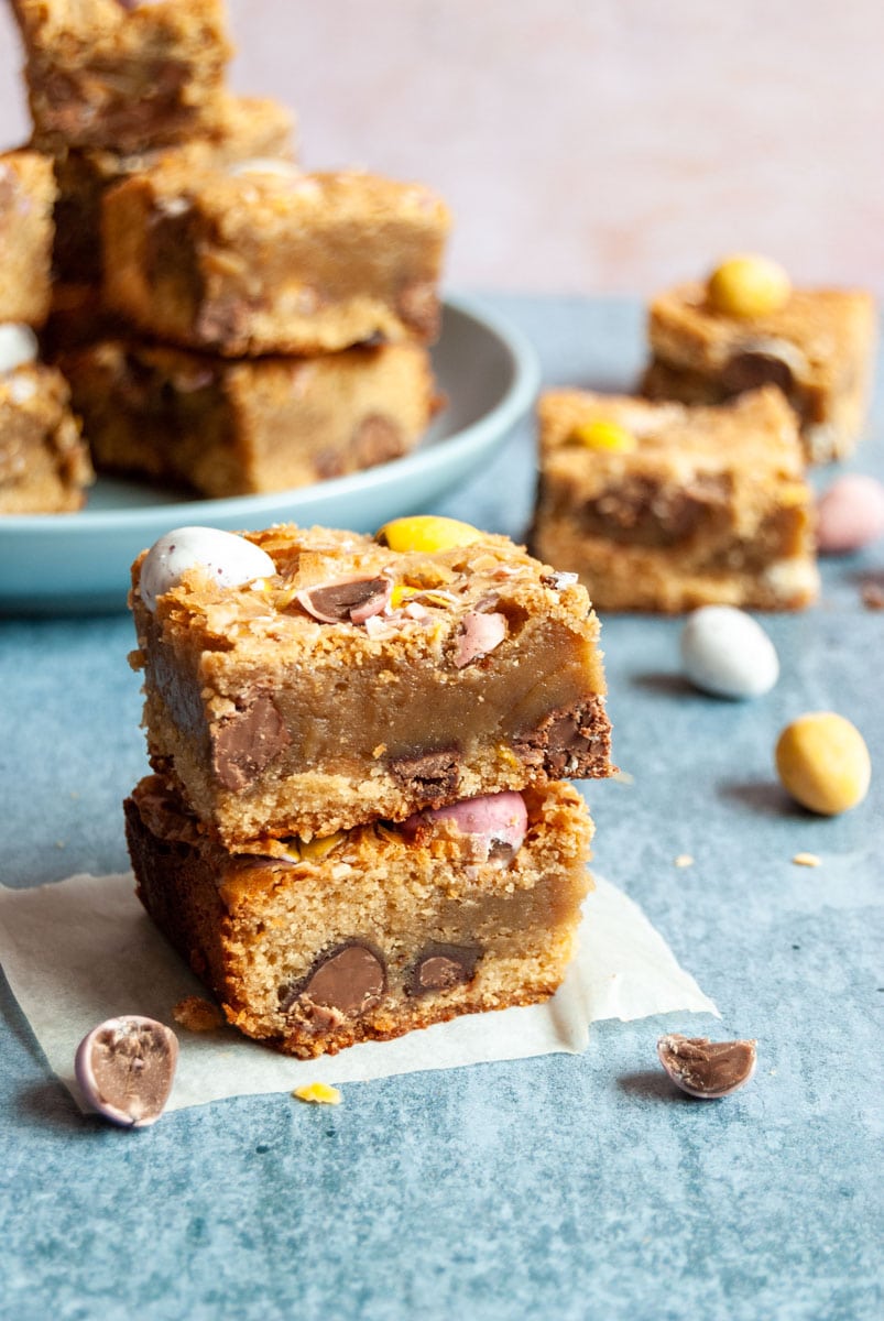 two blondies with mini coloured eggs stacked on top of each other on a piece of baking paper. More blondie bars can be partially seen on a blue plate. in the background.