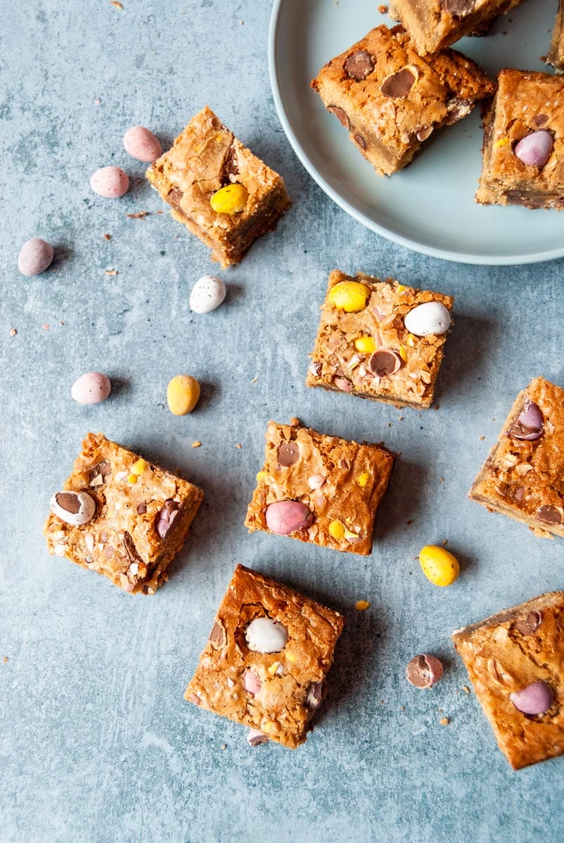 seven mini egg blondies bars on a blue background with pastel coloured mini eggs placed around the blondies.