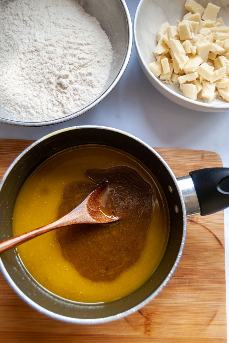 a small saucepan of melted butter and brown sugar with a wooden spoon, a silver bowl of flour and a white bowl of chopped white chocolate.