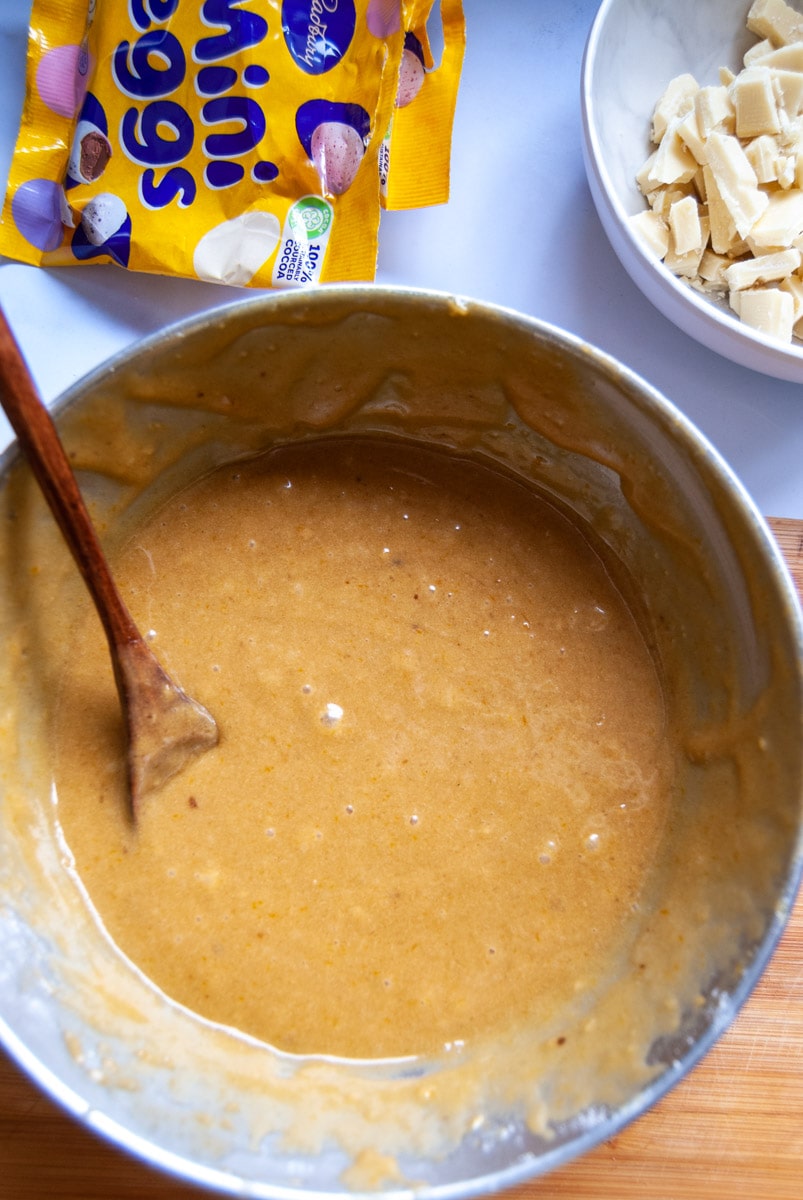 a silver mixing bowl of blondie batter with a wooden spoon, a packet of Cadbury mini eggs and a white bowl of chopped white chocolate.