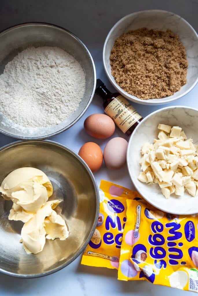 an overhead image of two packets of cadbury mini eggs, siliver bowls of butter and flour, white bowls of brown sugar and chopped white chocolate, three eggs and a bottle of vanilla extract.