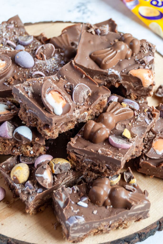 squares of chocolate fridge cake on a wooden board topped with mini creme eggs, Easter bunny chocolates and mini chocolate eggs in pastel shades.