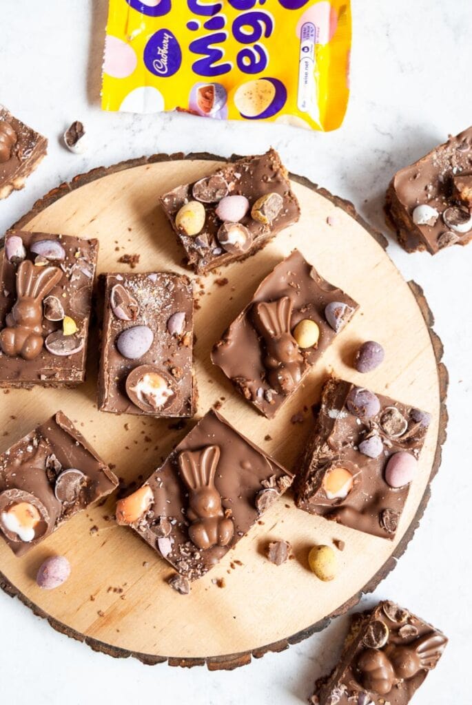 squares of chocolate fridge cake on a wooden board topped with Easter bunny chocolates, mini pastel coloured chocolate eggs and mini creme eggs.