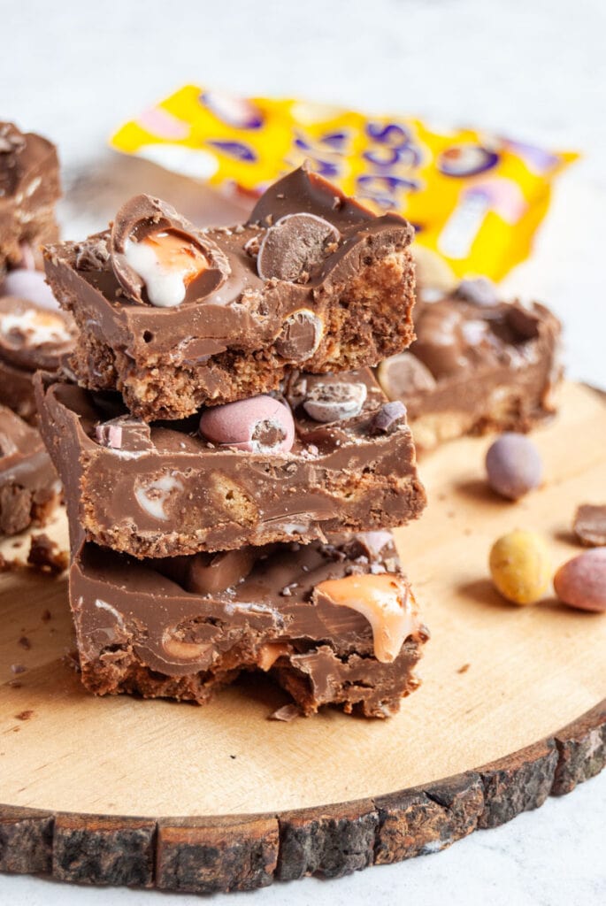 three slices of chocolate fridge cake stacked on top of each other with mini creme eggs and pastel coloured chocolate mini eggs sitting on a wooden board.