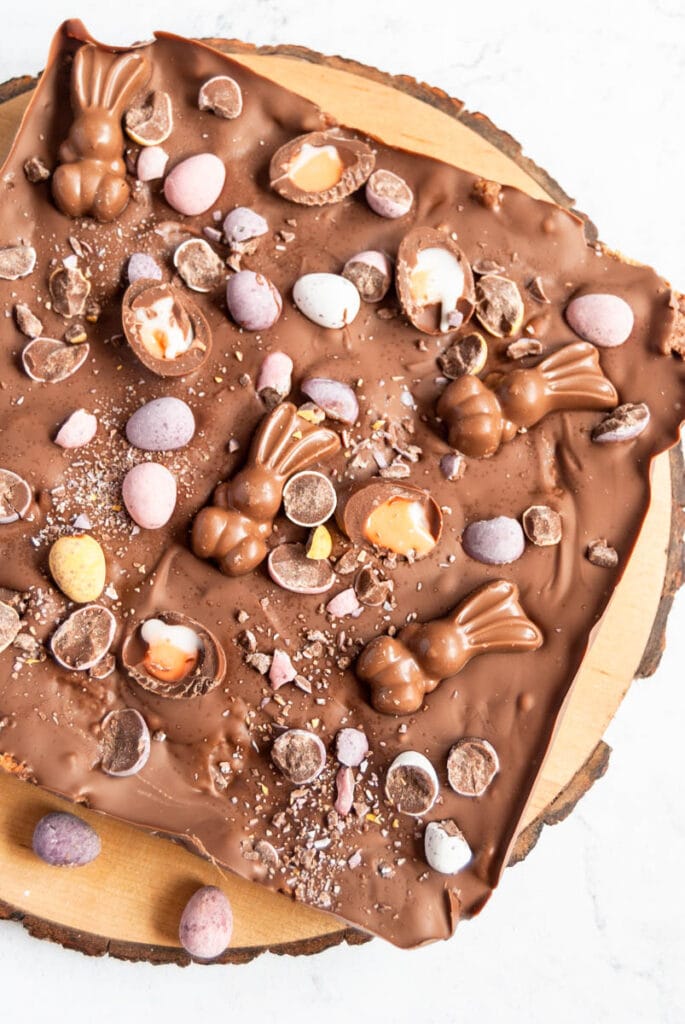 a uncut slab of chocolate fridge cake decorated with Easter bunny chocolates, mini eggs and mini creme eggs on a wooden board.