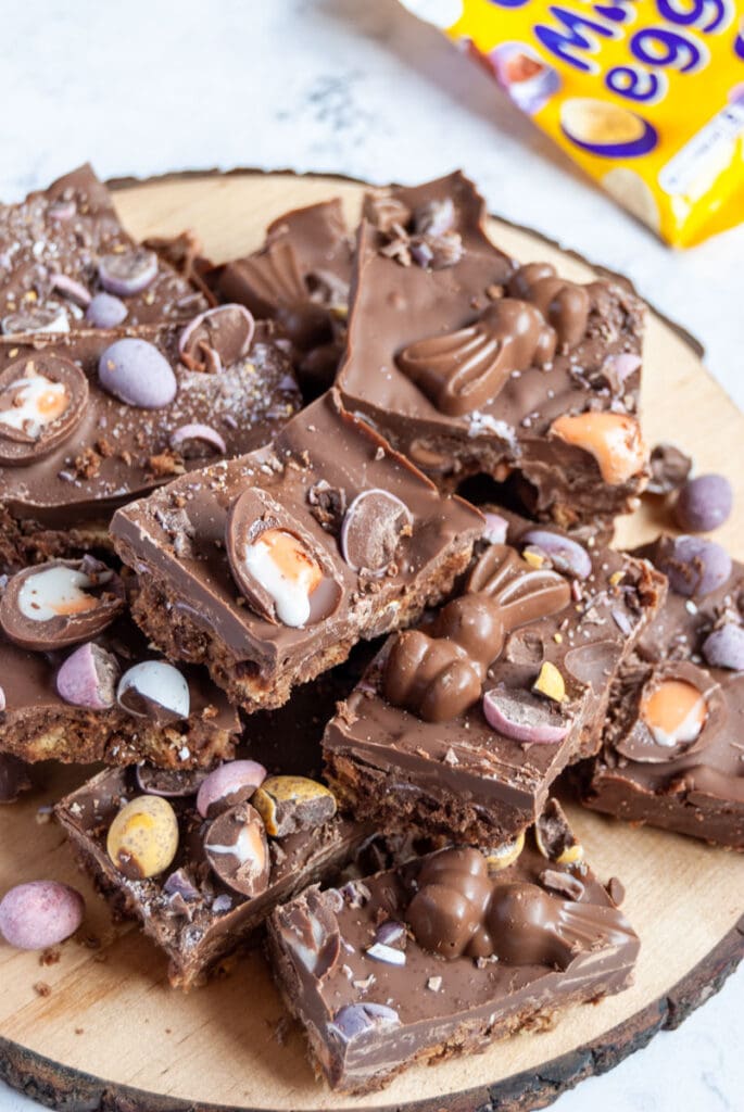 Squares of chocolate fridge cake topped with mini creme eggs, mini eggs and chocolate bunnines on a wooden board.