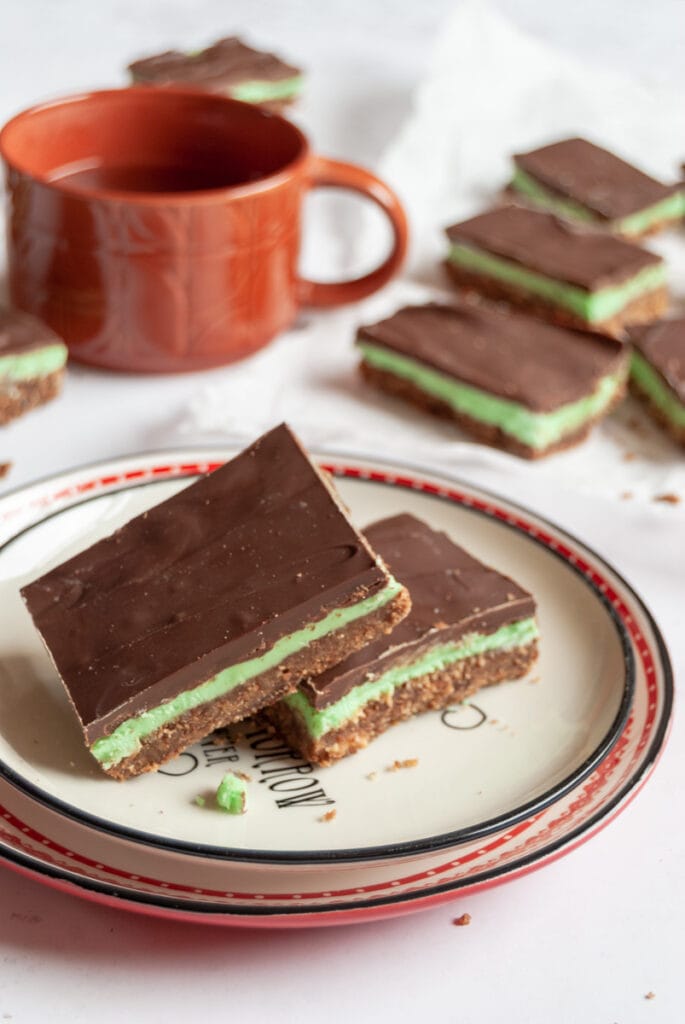 two slices of peppermint slice with a green fondant filling on a plate, an orange cup of tea and more peppermint slices in the background.