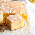 a lemon drizzle traybake cut into squares on a wire cooling rack lined with baking paper. A white plate with a square of lemon cake and fresh lemon slices are in the background.