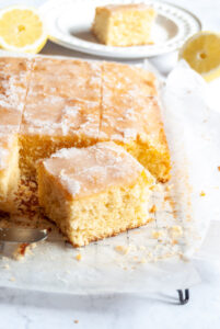 a lemon drizzle traybake cut into squares on a wire cooling rack lined with baking paper. A white plate with a square of lemon cake and fresh lemon slices are in the background.