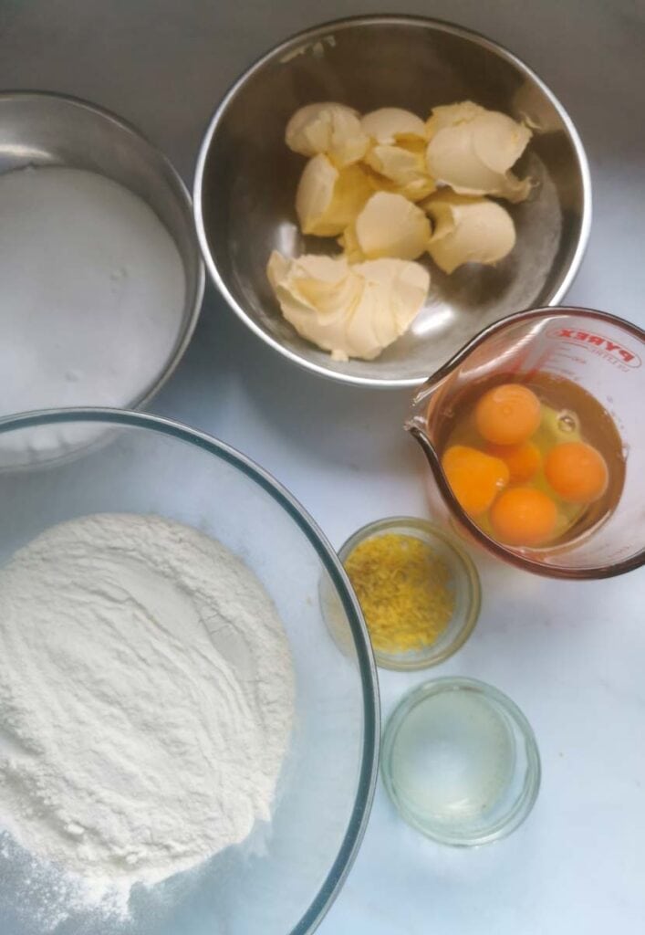 a glass mixing bowl of flour, silver bowls of butter and sugar, a jug of eggs and pots of lemon zest and juice.