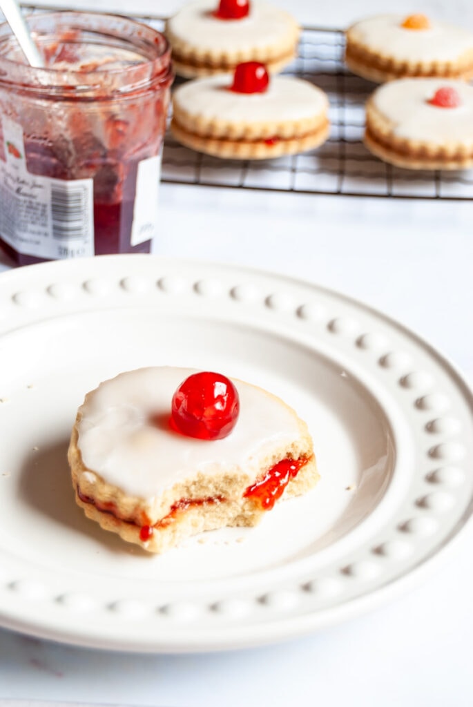 a jam filled sandwich biscuit topped with icing and a cherry with a bite taken out of it on a white plate