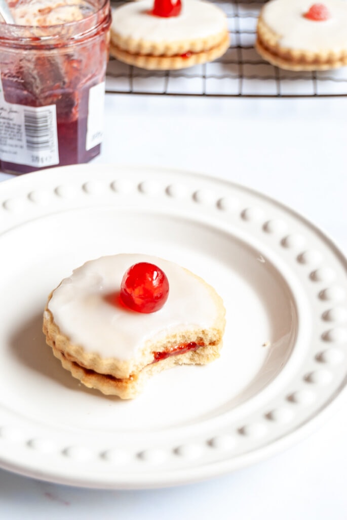 an jam filled biscuit topped with icing sugar and a glace cherry on a white plate.  A jar of strawberry jam and a wire rack with more biscuits sits in the background.