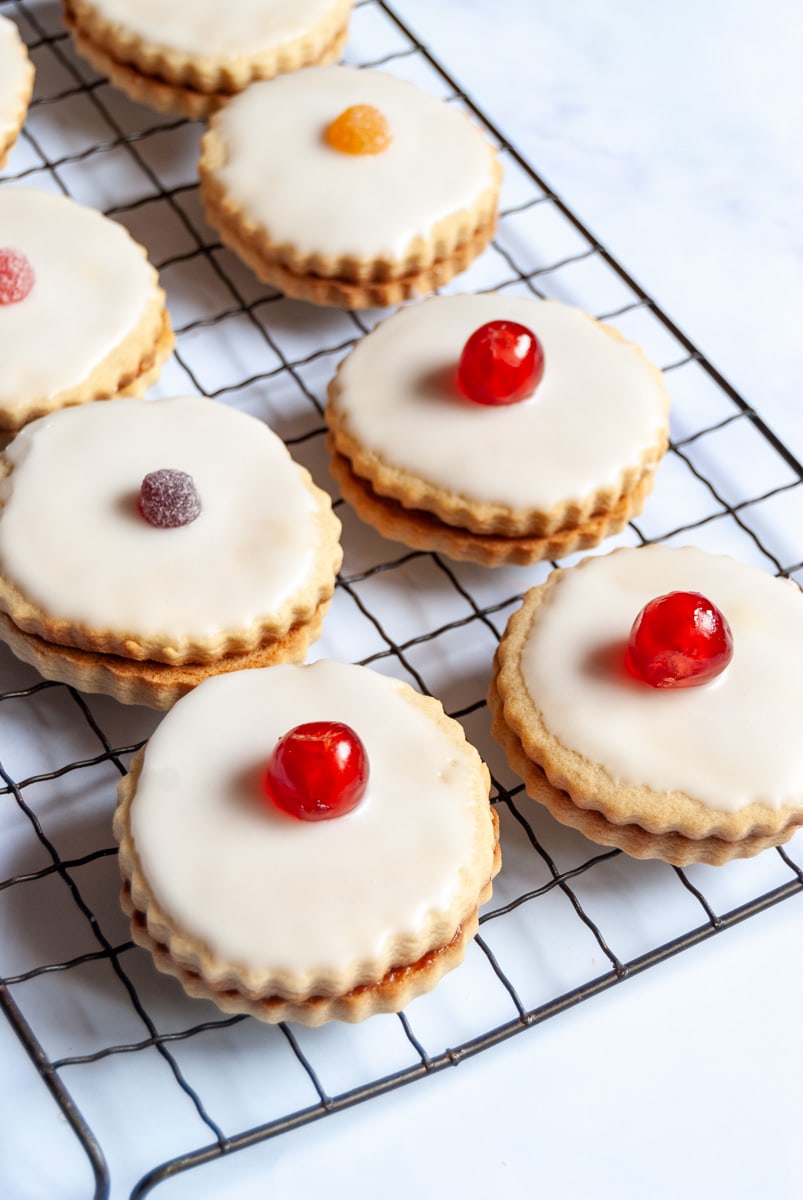 eight jam filled sandwich biscuits topped with white icing and a glace cherry or coloured sweet on a wire cooling rack.