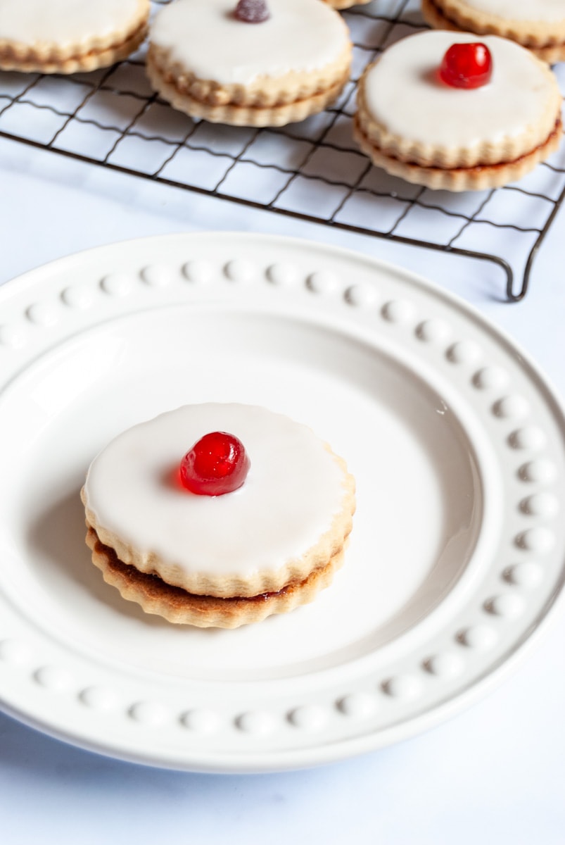 an Empire biscuit topped with icing and a glace cherry on a white plate.  A wire cooling rack with more biscuits sits behind the plate.