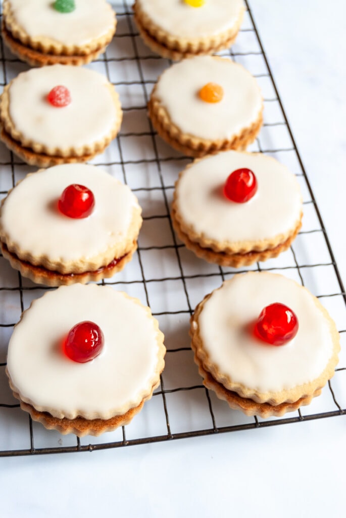eight sandwich biscuits filled with jam, topped with icing and decorated with a cherry on a wire cooling rack.