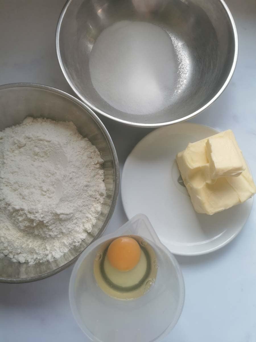 a large mixing bowl of flour, a bowl of sugar, a plate of butter and a small plastic jug with an egg cracked into it. 