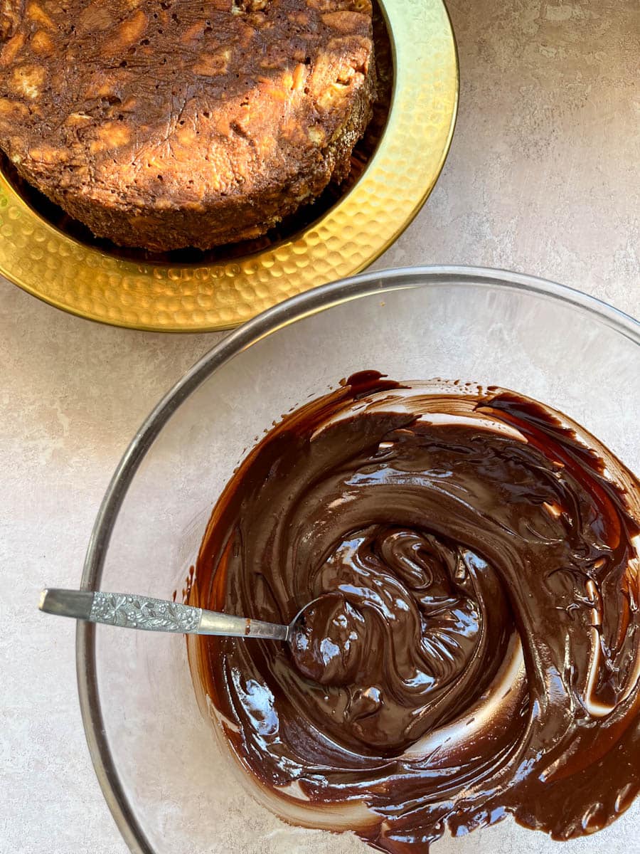 a large glass mixing bowl of chocolate ganache and around chocolate biscuit cake on a gold cake stand.