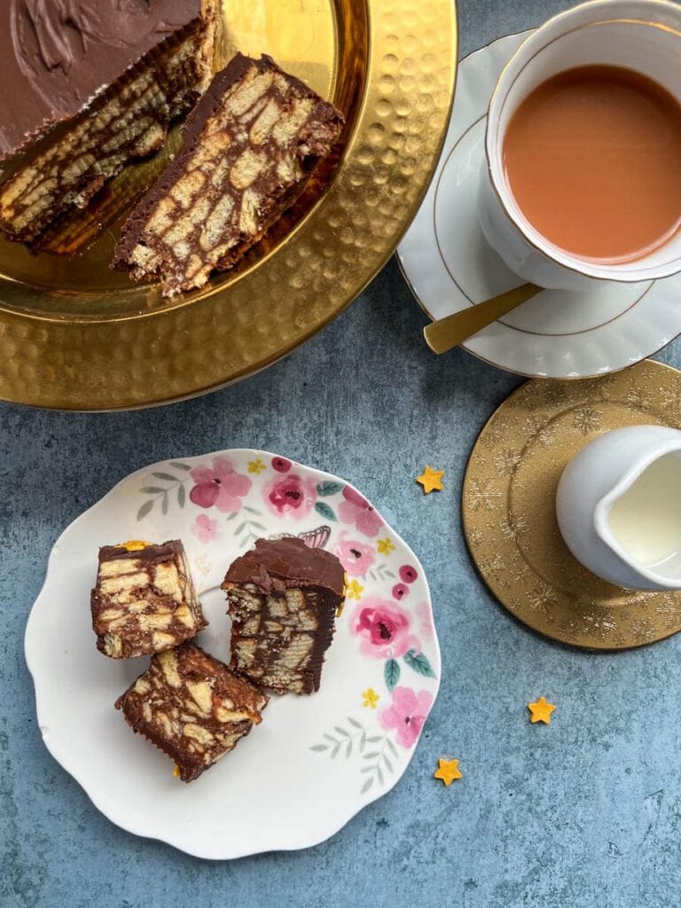 three pieces of chocolate biscuit cake on a floral plate, a white and gold cup of tea on a saucer and a small jug of milk sat on a gold coaster.