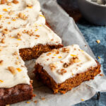 a large carrot cake with a slice cut out with cream cheese icing and sprinkled with chopped walnuts and orange zest on a piece of baking paper. A small grey bowl and an orange can be partially seen in the background.