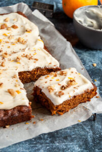 a large carrot cake with a slice cut out with cream cheese icing and sprinkled with chopped walnuts and orange zest on a piece of baking paper. A small grey bowl and an orange can be partially seen in the background.
