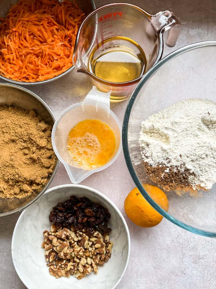various bowls filled with baking ingredients - brown sugar, flour and spices, chopped walnuts and sultanas, grated carrots, an orange and two measuring jugs of oil and beaten eggs.