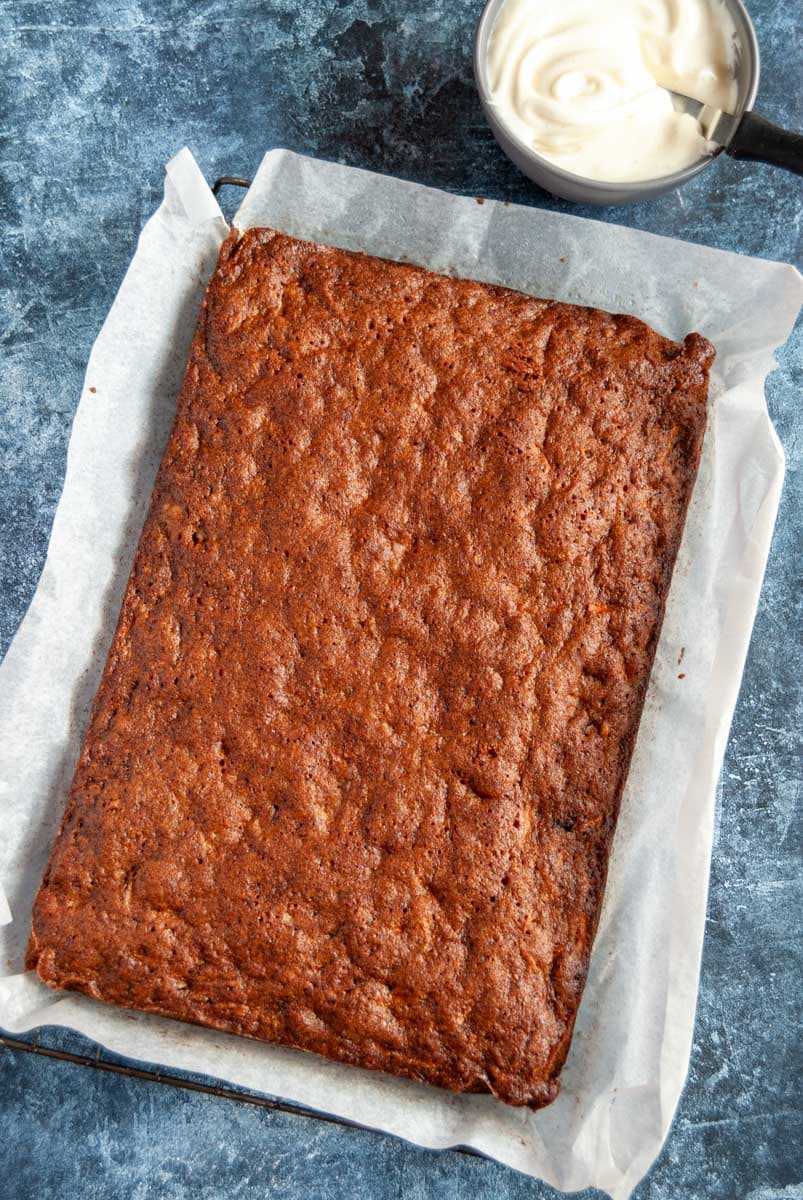 a large freshly baked carrot cake sitting on a wire cooling rack and a small grey bowl of cream cheese icing.